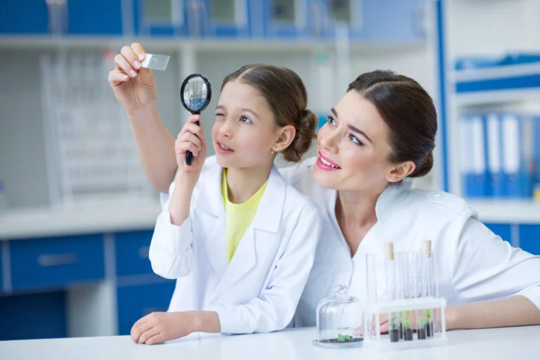 Woman teacher and girl student scientists looking at glass microscope slide through magnifier